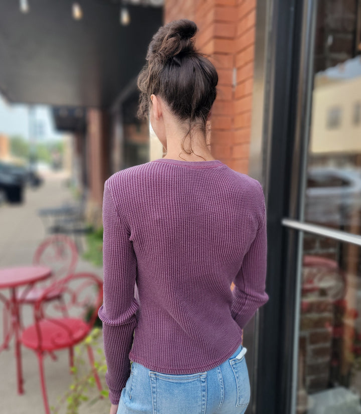 A person with dark hair in a bun, wearing the HYFVE, Inc Eliza Long Sleeve in purple and light blue jeans, stands outside by a brick wall and glass window, facing away from the camera. Red chairs and a table are visible nearby.