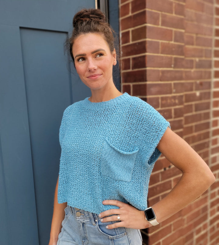 A woman with brown hair in a bun wears the HYFVE, Inc Sophia Short Sleeve Sweater in light blue, paired with denim shorts and a smartwatch. She stands by a blue door and brick wall, looking to the side with a soft smile.