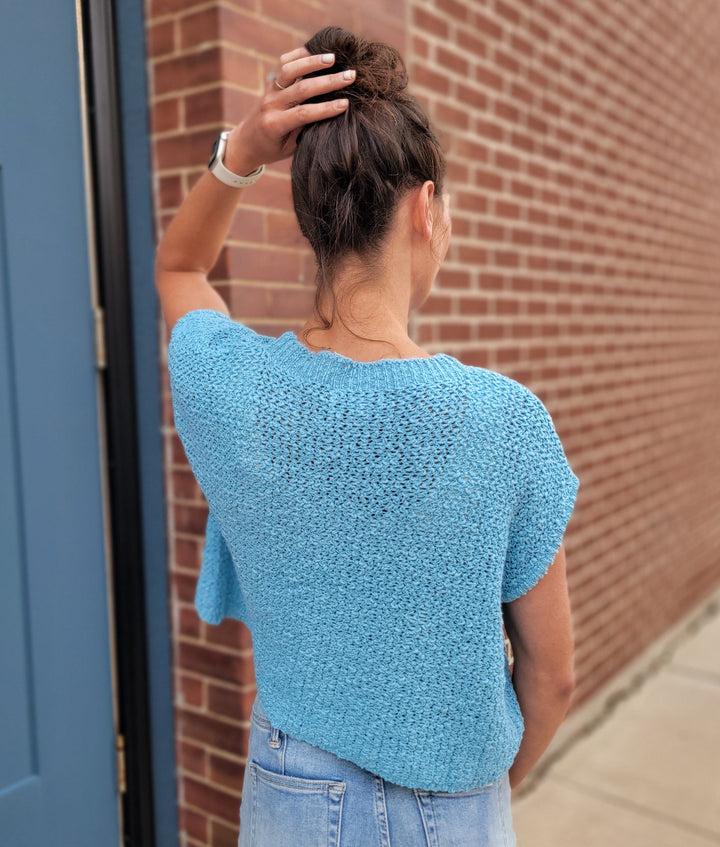 A woman faces a brick wall with her back to the camera, wearing the HYFVE, Inc Sophia Short Sleeve Sweater and light denim jeans, her hair in a bun and her left hand resting on the back of her head.
