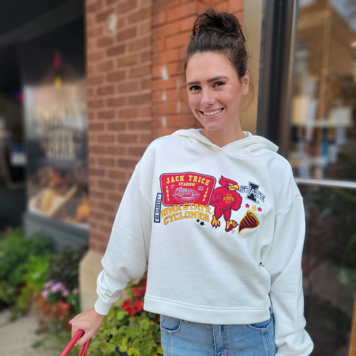 A smiling woman stands outside by a brick wall, wearing the Game Day Social ISU Cropped Hoodie featuring colorful graphics and patch emblems. She holds a red strap, with plants visible in the foreground.