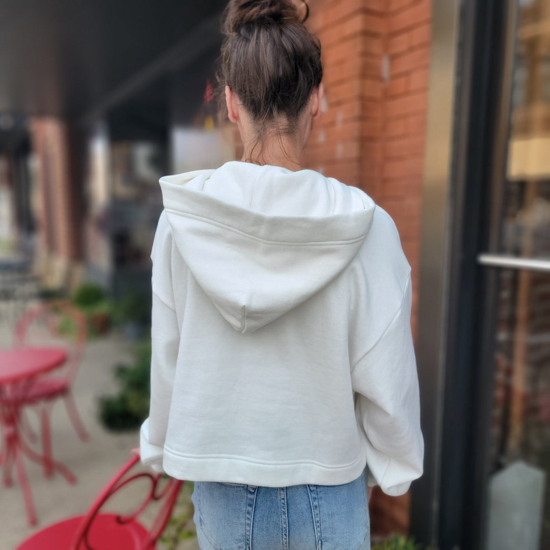 A woman with brown hair in a bun, seen from behind, wears the Game Day Social ISU Cropped Hoodie and blue jeans near a red chair and table outside a brick building with glass windows.
