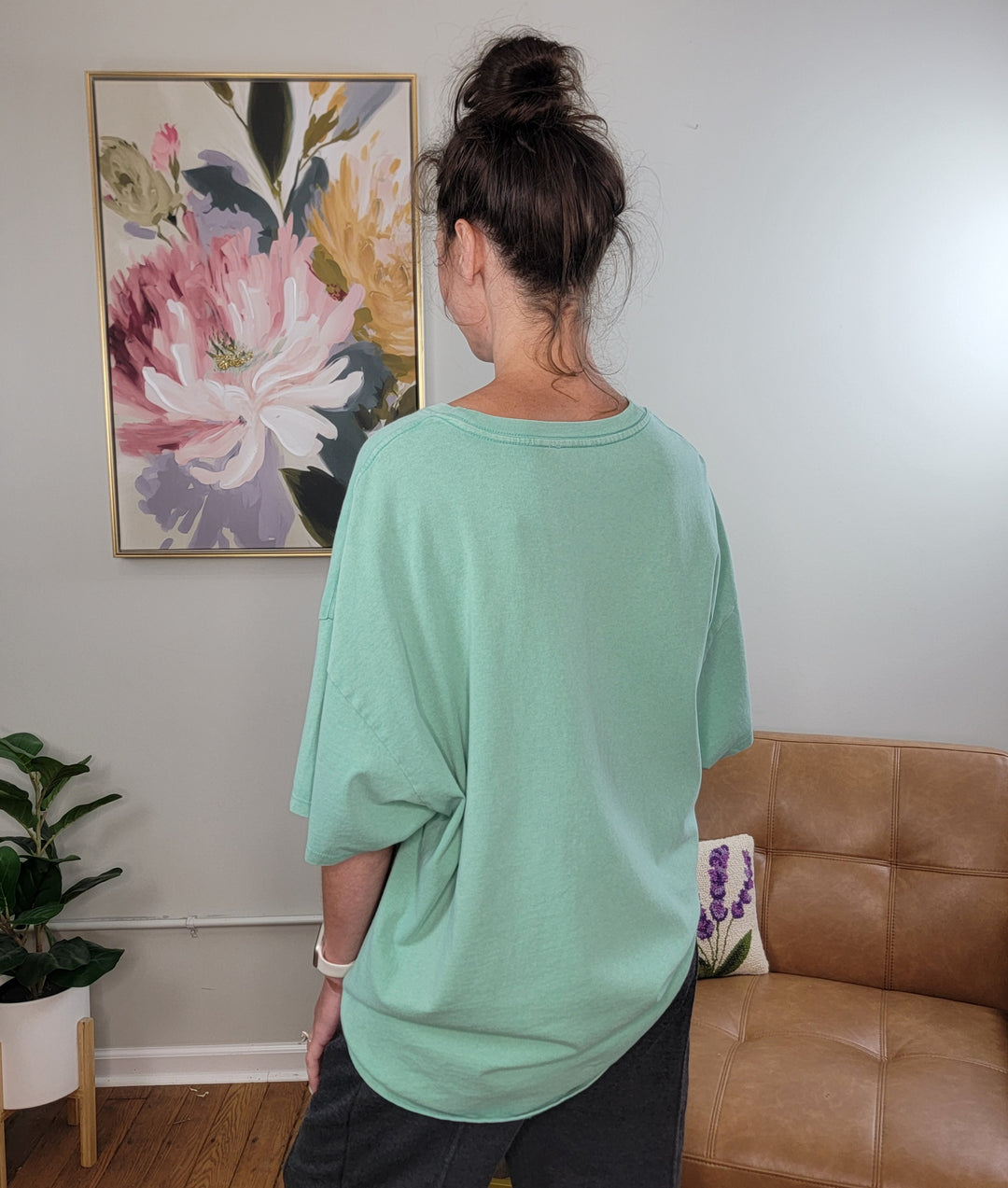 A person with dark hair in a messy bun, wearing the HYFVE, Inc Presley Oversized Tee in emerald and dark pants, stands with their back to the camera in a cozy room featuring a floral painting, brown couch, and potted plant.