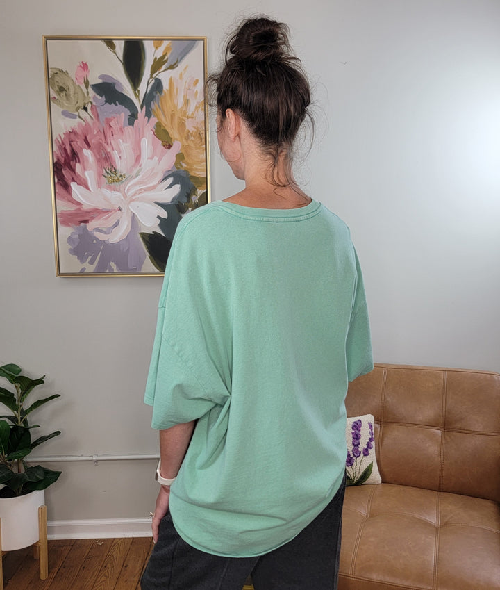 A person with dark hair in a messy bun, wearing the HYFVE, Inc Presley Oversized Tee in emerald and dark pants, stands with their back to the camera in a cozy room featuring a floral painting, brown couch, and potted plant.