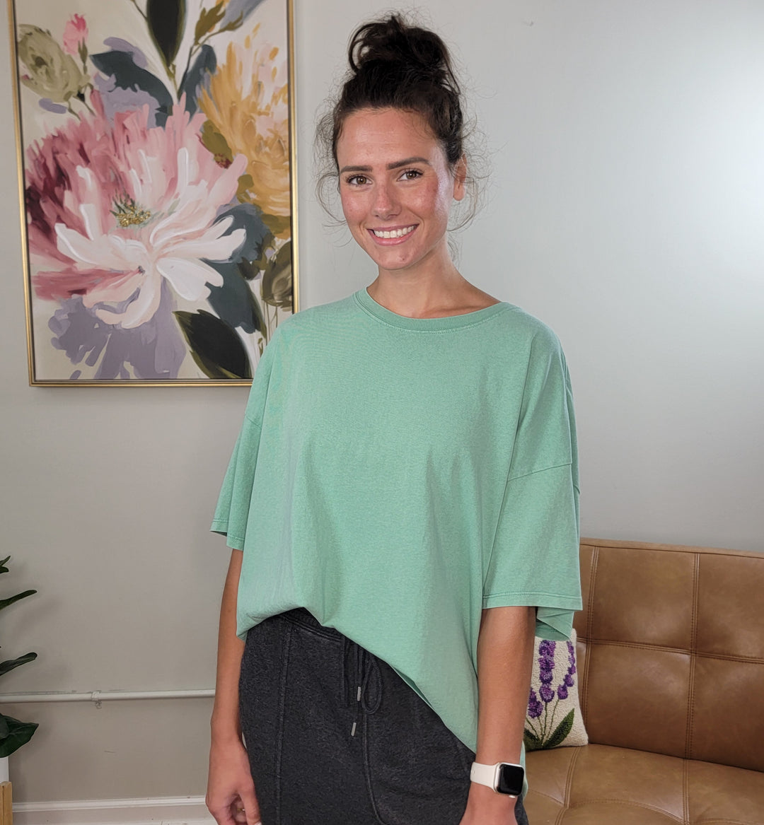 A woman with dark hair in a bun wears the HYFVE, Inc Presley Oversized Tee in emerald and dark pants, smiling in a living room decorated with a floral painting and tan couch.