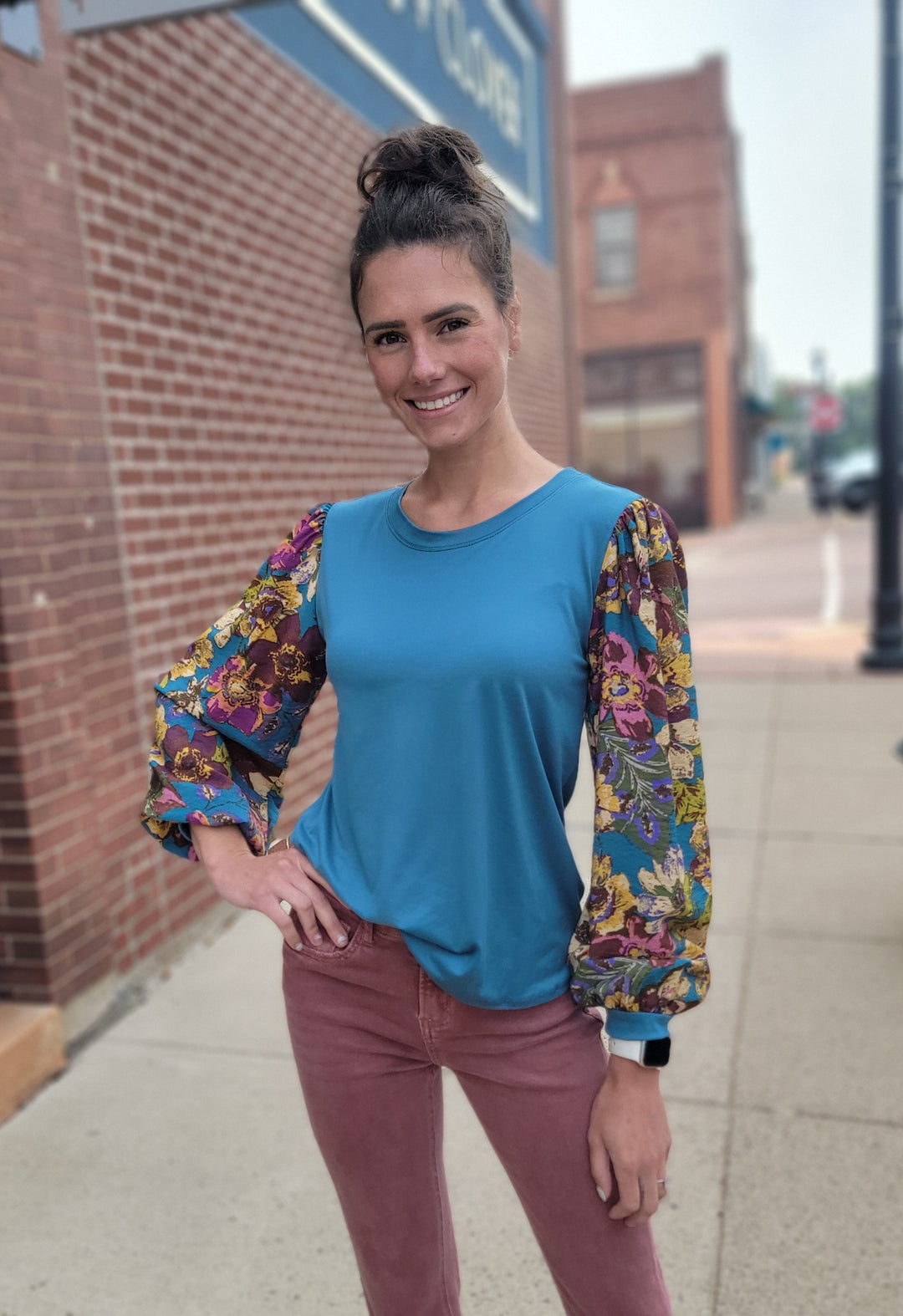 A woman with brown hair in a bun stands on a sidewalk in front of a brick building, smiling and wearing the Les Amis Natalie Floral Sleeve Blouse in turquoise with mauve pants on a cloudy day.