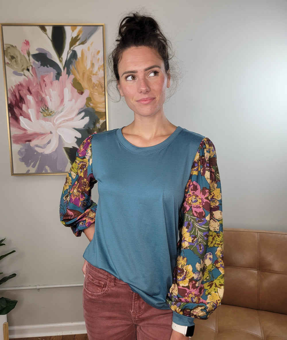 A woman with dark hair in a bun stands indoors, wearing the Les Amis Natalie Floral Sleeve Blouse—a turquoise top with lightweight, stretchy floral print balloon sleeves—and maroon pants. She looks to the side before a floral painting and beige wall.