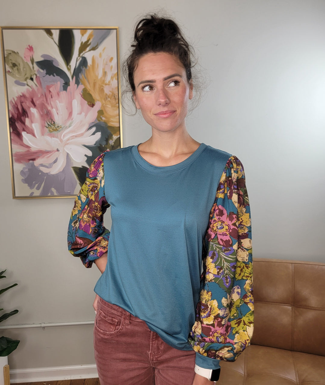 A woman with dark hair in a bun stands indoors, wearing the Les Amis Natalie Floral Sleeve Blouse—a turquoise top with lightweight, stretchy floral print balloon sleeves—and maroon pants. She looks to the side before a floral painting and beige wall.