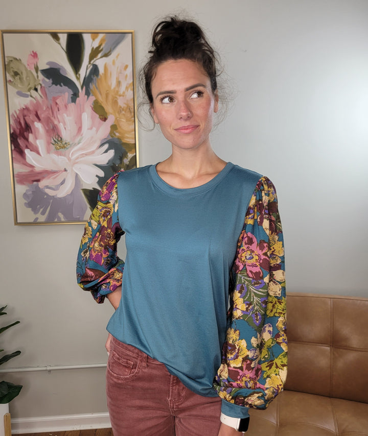 A woman with dark hair in a bun stands indoors, wearing the Les Amis Natalie Floral Sleeve Blouse—a turquoise top with lightweight, stretchy floral print balloon sleeves—and maroon pants. She looks to the side before a floral painting and beige wall.