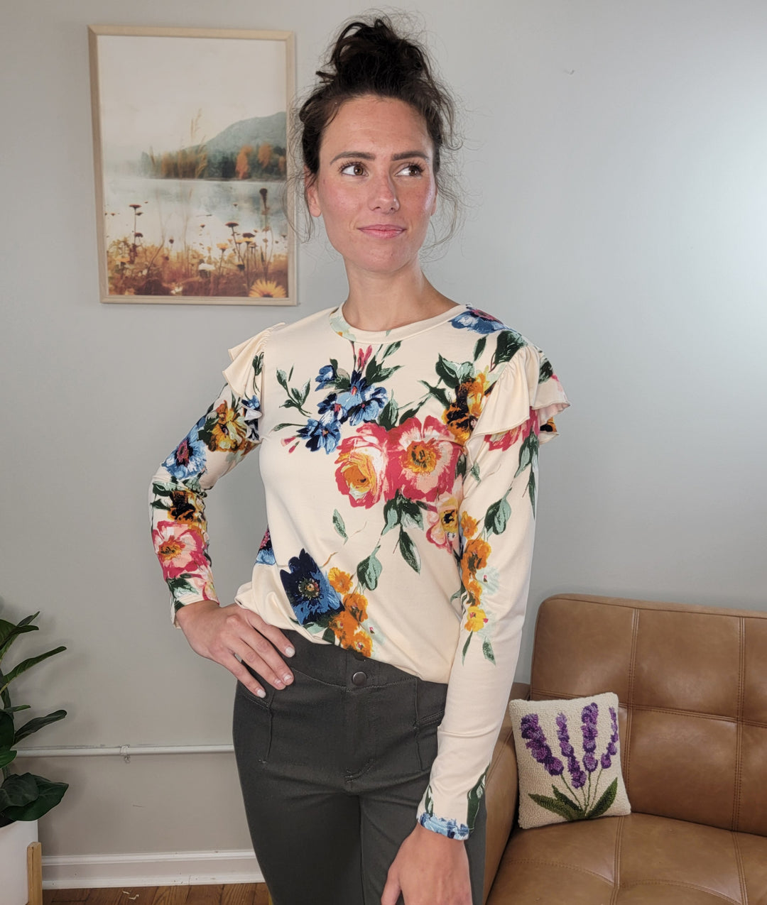 A woman wears the Les Amis Billie Floral Long Sleeve top with dark pants, standing indoors near a framed artwork, a tan sofa, and a lavender-patterned pillow while gazing to the side.