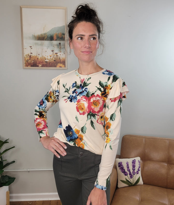 A woman wears the Les Amis Billie Floral Long Sleeve top with dark pants, standing indoors near a framed artwork, a tan sofa, and a lavender-patterned pillow while gazing to the side.