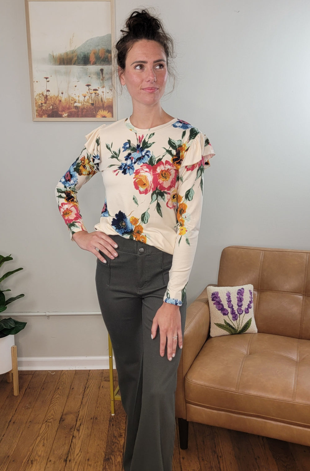 A woman with dark hair in a messy bun stands indoors near a tan armchair and nature-themed wall art, wearing the Les Amis Billie Floral Long Sleeve top and dark wide-leg pants.