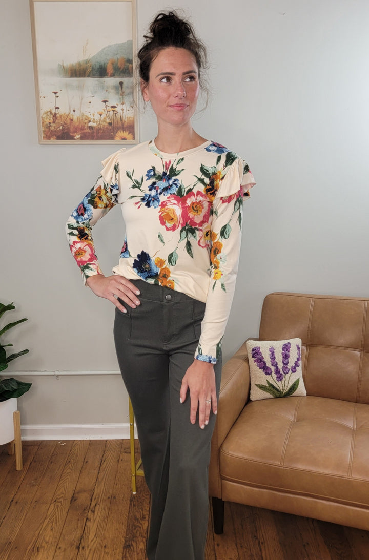 A woman with dark hair in a messy bun stands indoors near a tan armchair and nature-themed wall art, wearing the Les Amis Billie Floral Long Sleeve top and dark wide-leg pants.