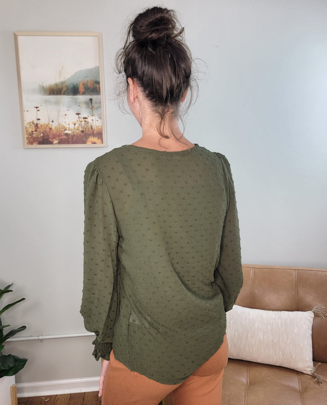 A woman with dark hair in a bun stands indoors, wearing the Les Amis Adeline Long Sleeve Blouse in olive and light brown pants. A framed nature picture hangs on the wall, and a tan couch is nearby.
