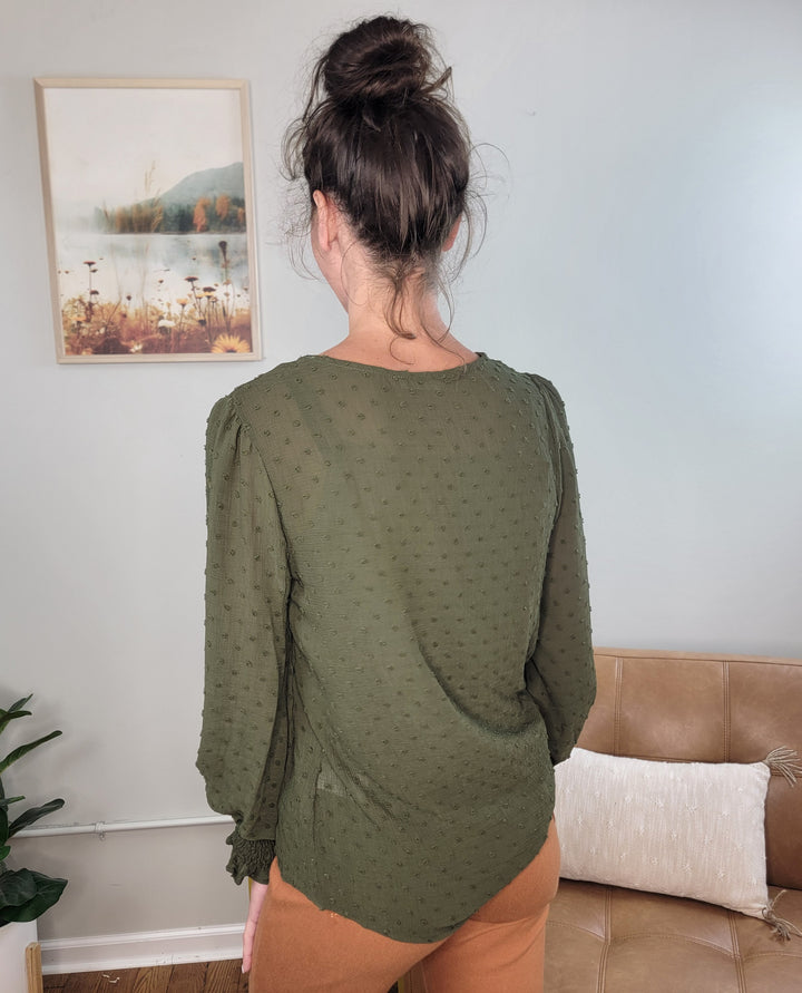 A woman with dark hair in a bun stands indoors, wearing the Les Amis Adeline Long Sleeve Blouse in olive and light brown pants. A framed nature picture hangs on the wall, and a tan couch is nearby.