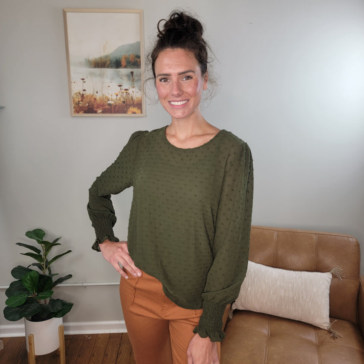 A woman with brown hair in a bun smiles in a cozy room, wearing the Les Amis Adeline Long Sleeve Blouse and brown pants. The space features a tan sofa, a plant, and a nature-themed picture on the wall.
