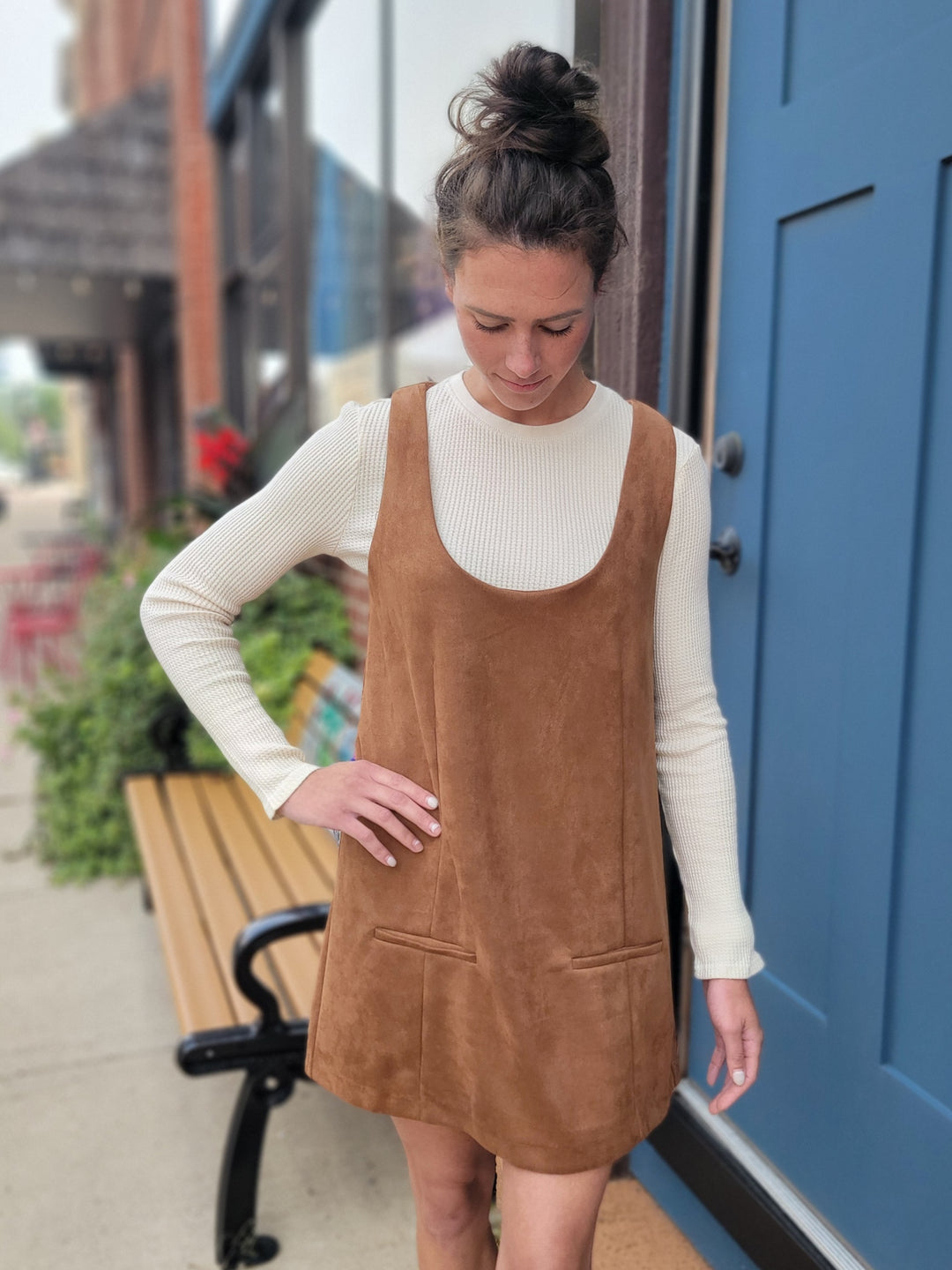 A woman stands outside by a blue door and bench, wearing the HYFVE, Inc Isabell Dress—a brown suede shift with pockets—over a cream long-sleeve top. Her hair is in a bun as nearby plants and buildings create a charming fall vibe.