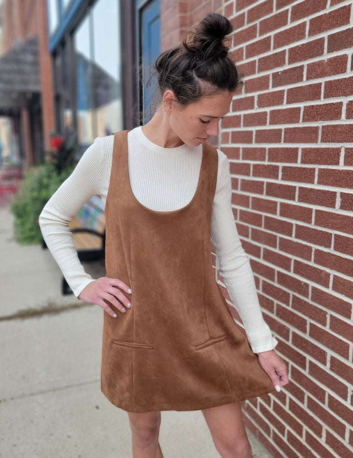 A woman with dark hair in a bun wears the Isabell Dress by HYFVE, Inc—a brown suede shift—over a cream long-sleeve top, standing by a red brick wall on a city sidewalk, looking down and touching her chic fall outfit.