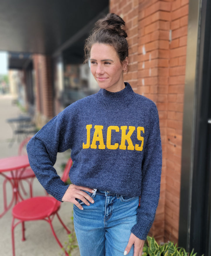 A woman stands outdoors by a brick wall in the Game Day Social Jacks Sweater, styled with blue jeans. Red chairs and tables are in the background, highlighting this cozy pick for chilly game days.