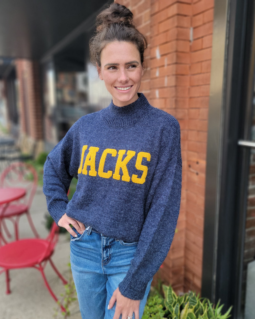 A woman with brown hair in a bun smiles at the camera while standing outside near red chairs and a brick wall, wearing a Game Day Social Jacks Sweater in blue with yellow letters and blue jeans.