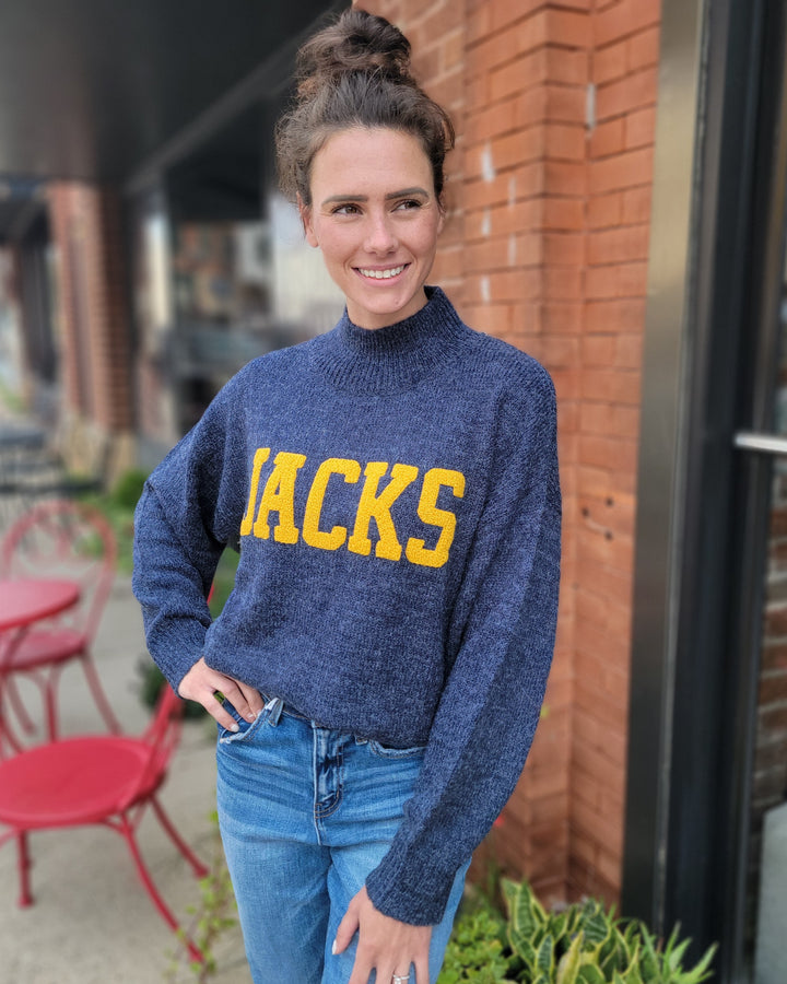 A woman with brown hair in a bun smiles at the camera while standing outside near red chairs and a brick wall, wearing a Game Day Social Jacks Sweater in blue with yellow letters and blue jeans.