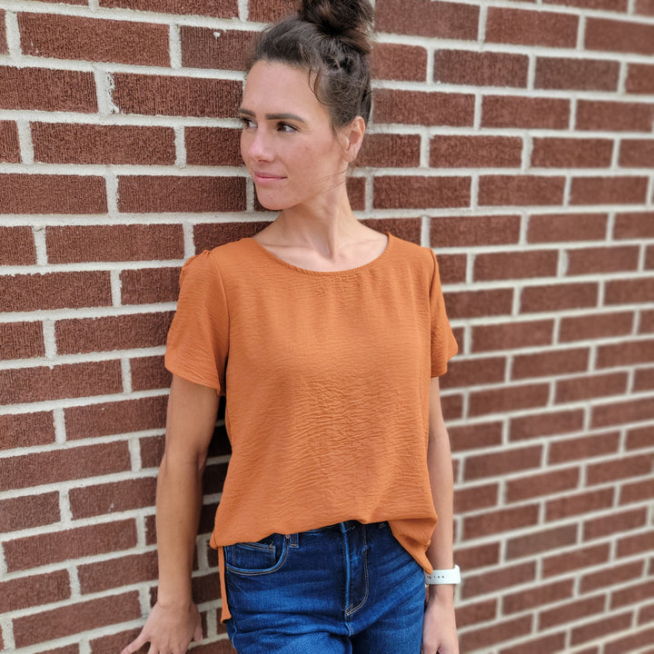 A woman in the Les Amis Norah Top stands against a brick wall, looking left. She pairs the rust-colored tulip sleeve top with blue jeans, wears her hair in a bun, and has a white smartwatch on her left wrist.