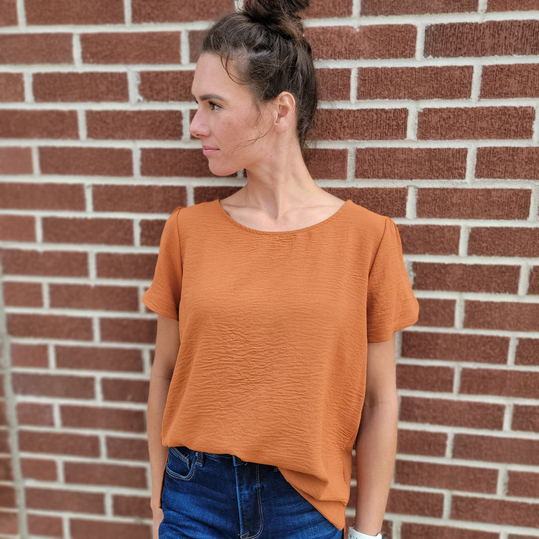 A woman with brown hair in a bun wears the Les Amis Norah Top and dark blue jeans, standing against a red brick wall with one hand in her pocket—a simple yet chic fall look.