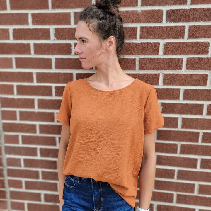 A woman with brown hair in a bun wears the Les Amis Norah Top and dark blue jeans, standing against a red brick wall with one hand in her pocket—a simple yet chic fall look.