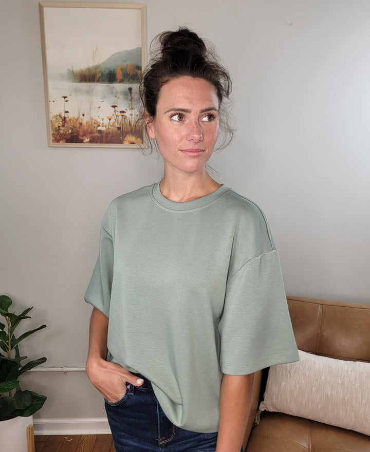 A woman with brown hair in a messy bun wears the HYFVE, Inc Emersyn Tee—a loose, light green crew neck top made from lightweight scuba fabric—paired with jeans. She stands indoors by a plant and tan couch, with a nature picture on the wall behind her.