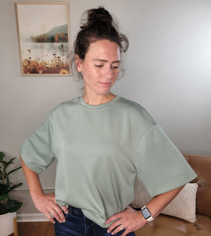 A woman with brown hair in a messy bun wears the light green Emersyn Tee by HYFVE, Inc—an oversized women's top in lightweight scuba material—with dark jeans. She stands indoors, hands on hips, looking down; artwork and a plant are behind her.
