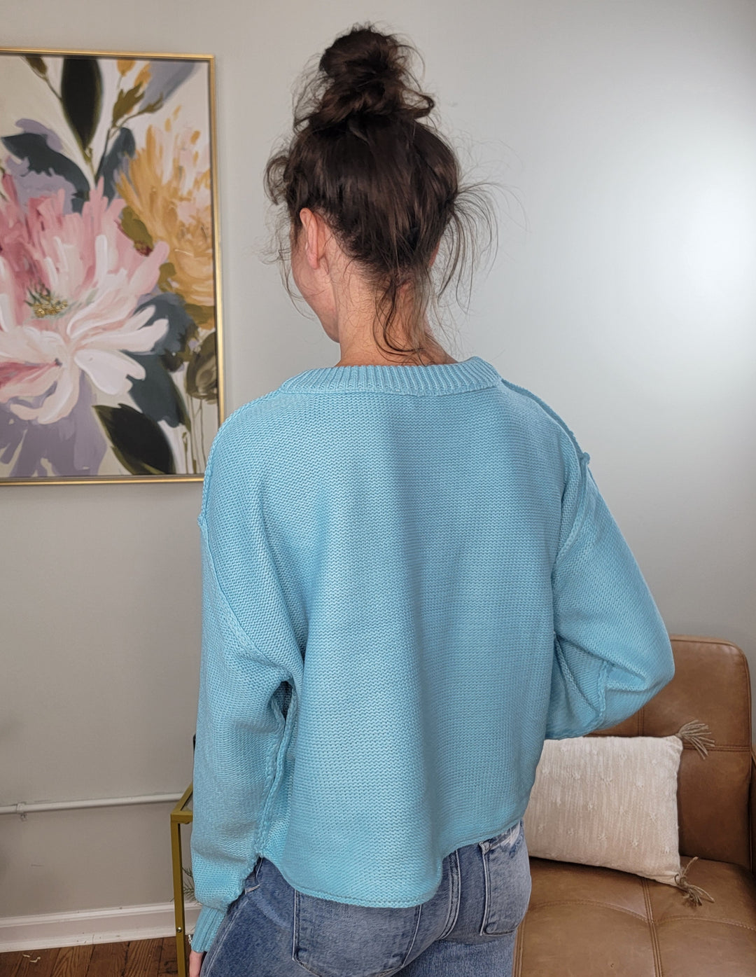 A person with dark hair in a messy bun stands with their back to the camera, wearing the HYFVE, Inc Nova Sweater in sky blue and blue jeans, in a room with light walls, a floral painting, and a tan couch.