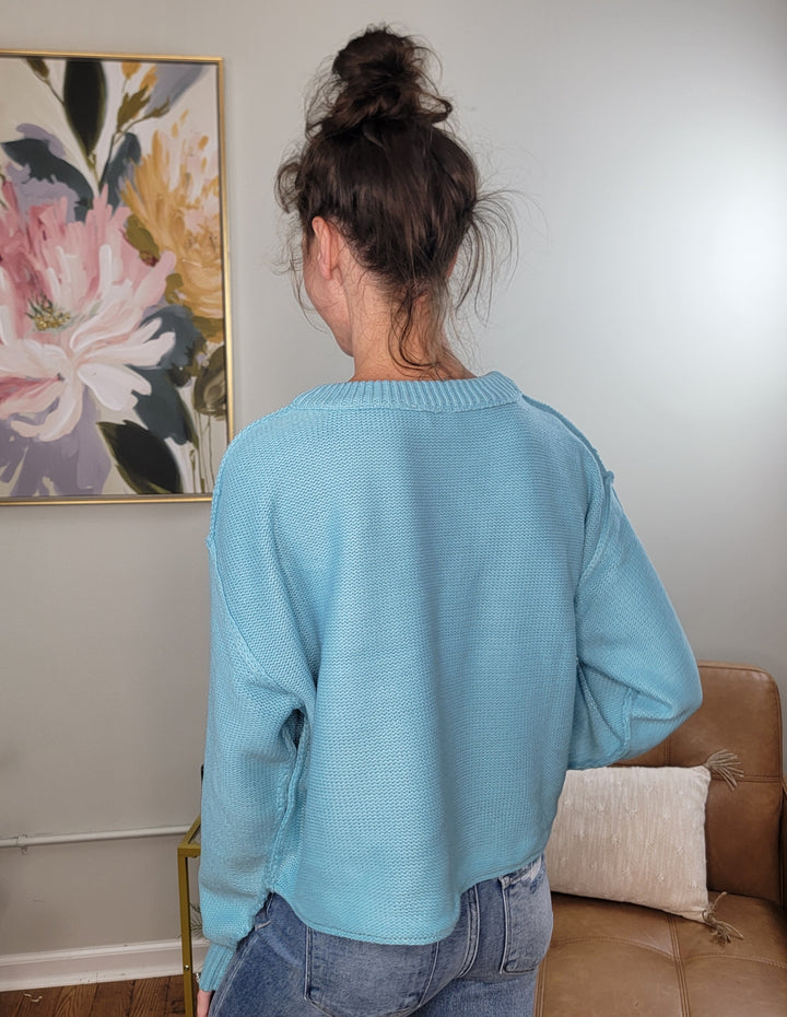 A person with dark hair in a messy bun stands with their back to the camera, wearing the HYFVE, Inc Nova Sweater in sky blue and blue jeans, in a room with light walls, a floral painting, and a tan couch.