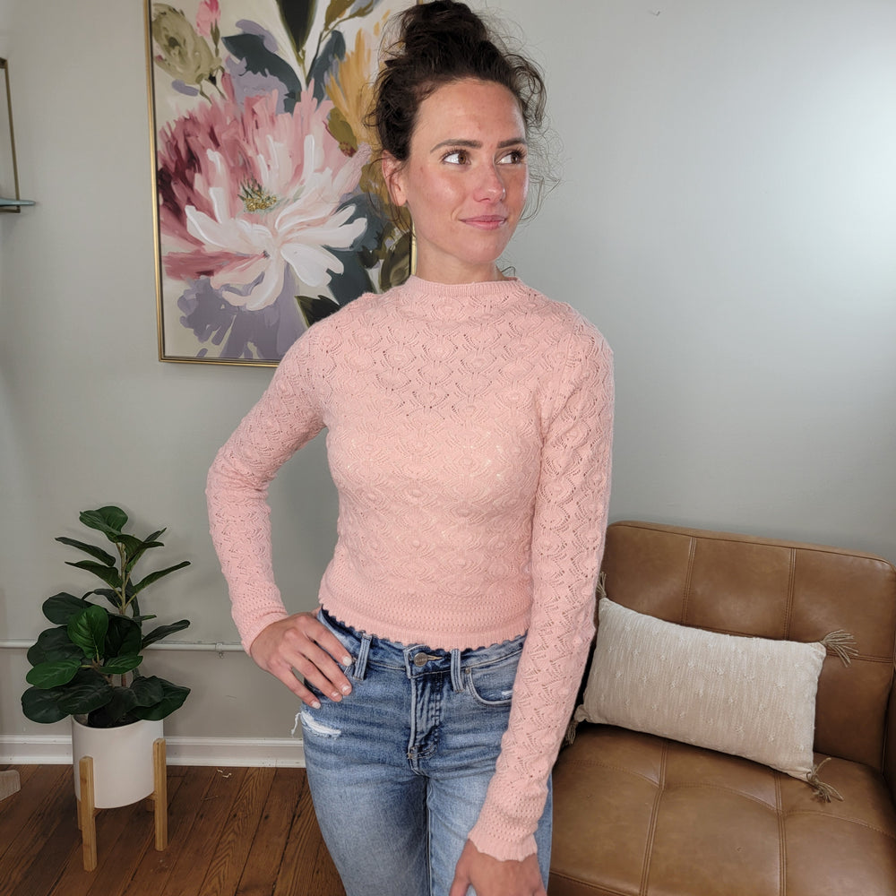 A woman with brown hair in a bun wears the light pink Pollie Sweater by HYFVE, Inc and blue jeans, standing indoors beside a tan chair and green plant, with a floral painting on the wall behind her.