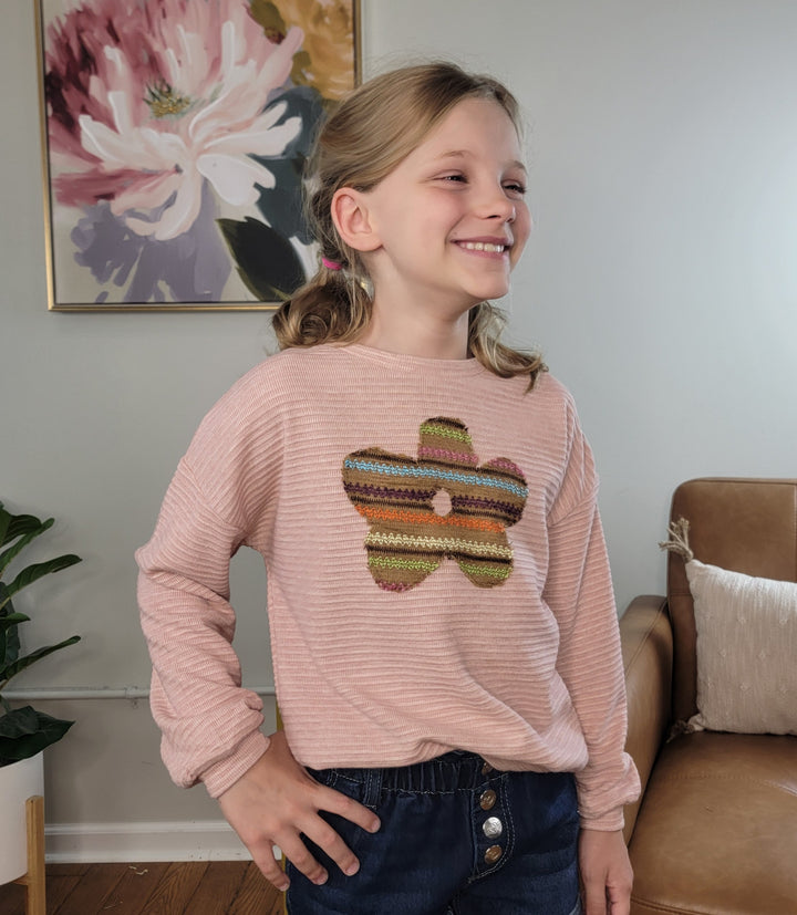 A smiling young girl stands indoors wearing the Hayden Girls Anika Flower Long Sleeve top and blue jeans. The light pink ribbed top features a colorful flower embellishment. A floral painting, plant, and tan chair are in the background.