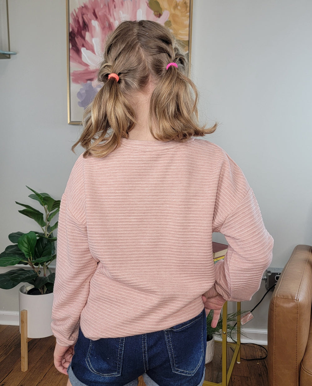 A girl with shoulder-length light brown hair in pigtails wears the Hayden Girls Anika Flower Long Sleeve, standing indoors with plants and a floral painting in the background, facing away from the camera.