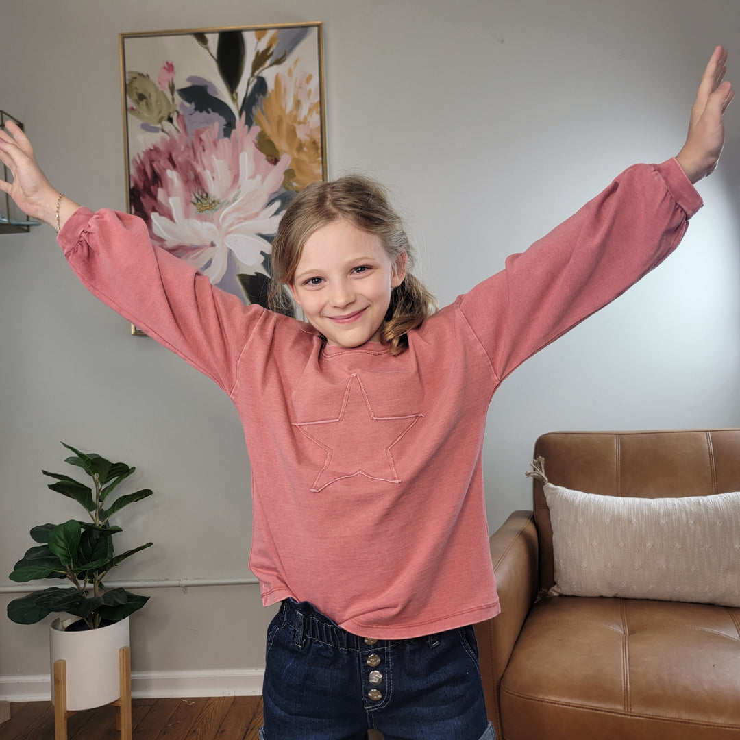 A smiling young girl with blonde hair wears the Hayden Girls Jaylee Star Crewneck and denim shorts indoors, arms outstretched. Behind her are a plant, armchair, and a floral painting.
