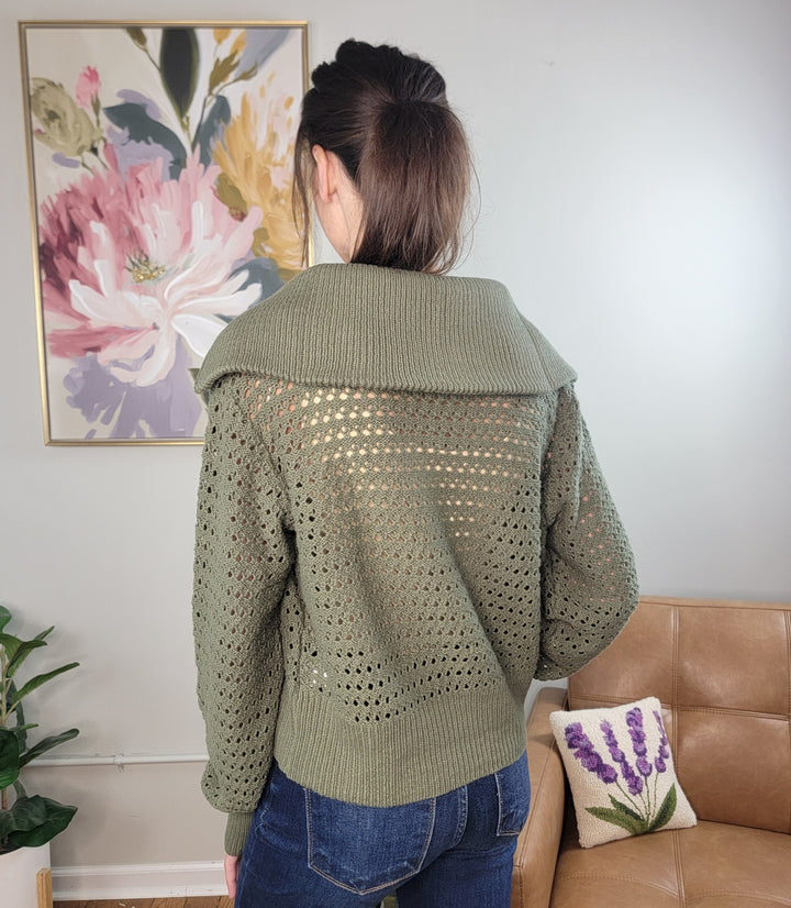 A woman with brown hair in a ponytail wears the Doe &amp; Rae Neriah Zip Front Knit Sweater with Fold Over Collar in olive green and blue jeans, standing with her back to the camera in a living room featuring a floral painting and tan couch.