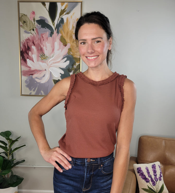 A woman with dark hair in a ponytail smiles indoors, wearing the Doe &amp; Rae Wrenlee Ruffle Detail Knit Tank Top in terra cotta and jeans. She stands before a floral painting, a plant, and a brown sofa with a lavender-patterned pillow—ideal for fall layering.