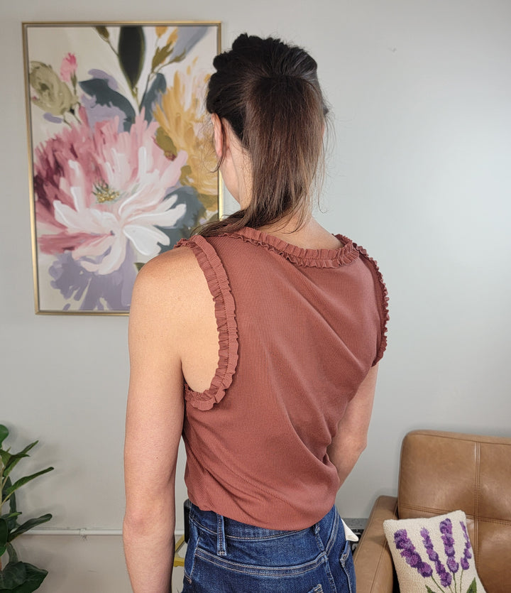 A woman with long brown hair in a ponytail stands with her back to the camera, wearing the Doe &amp; Rae Wrenlee Ruffle Detail Knit Tank Top in terra cotta and blue jeans. Indoors, a floral painting and beige couch are visible. Perfect for fall layering.