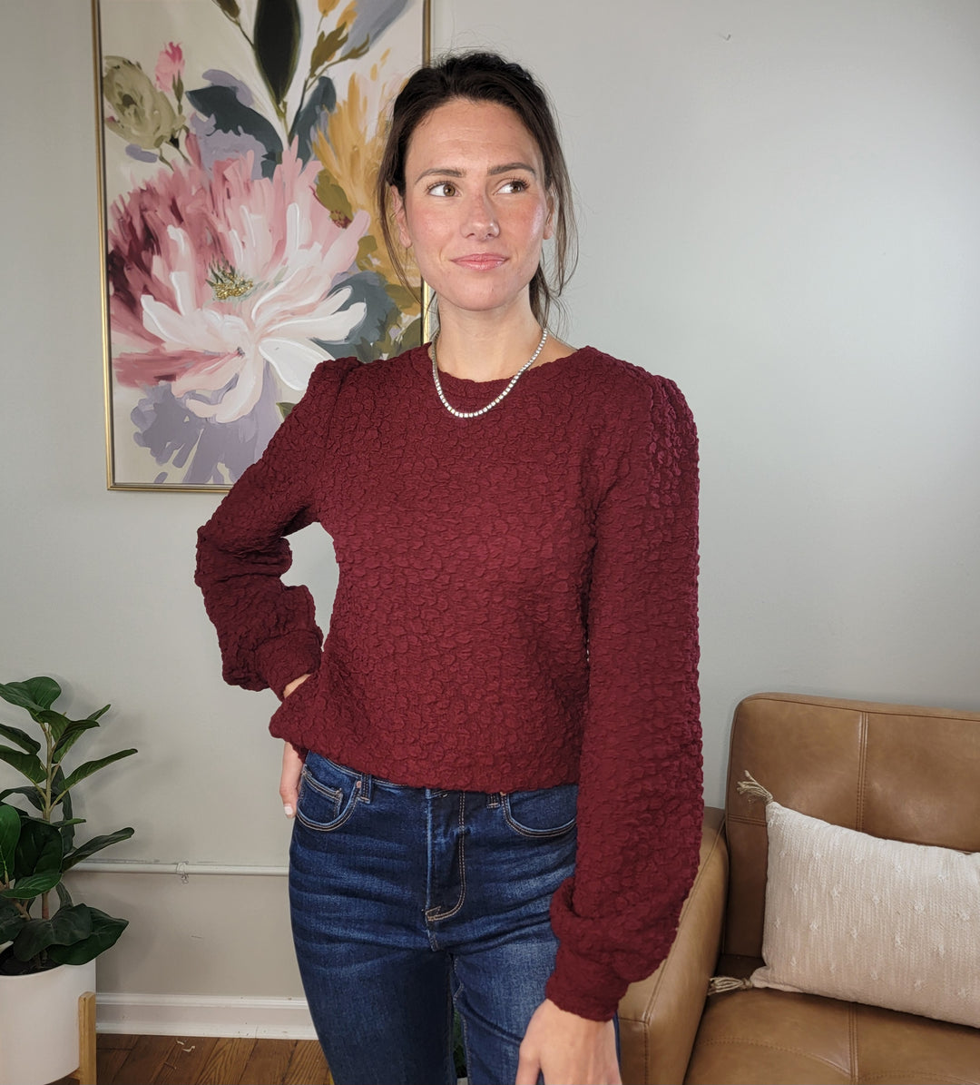 A woman stands indoors near a tan couch and potted plant, wearing the Doe & Rae Amayah Top in burgundy, blue jeans, and a pearl necklace, with a large floral painting on the wall behind her.