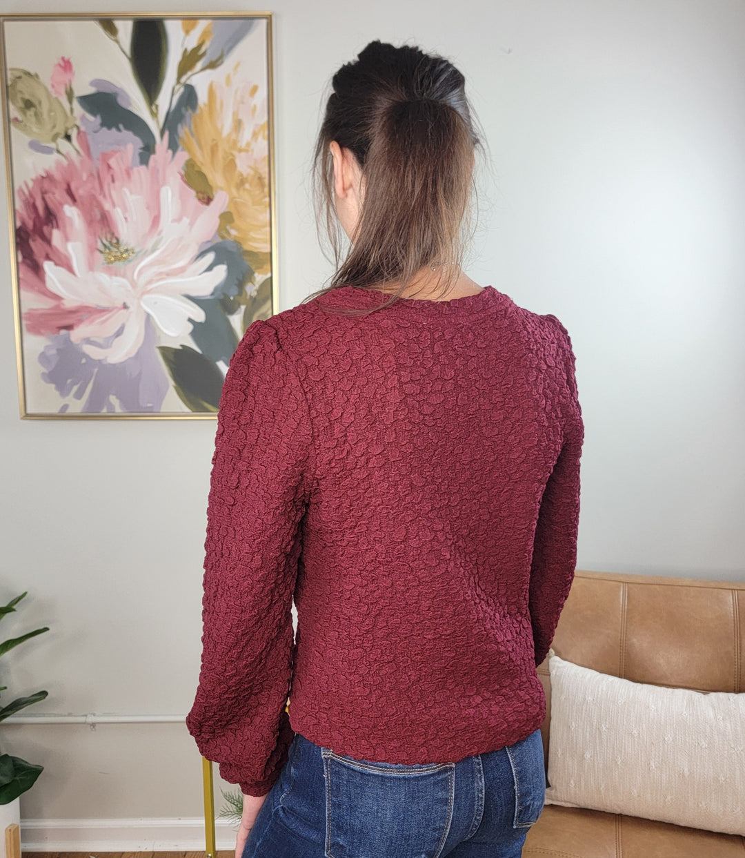 A woman with long brown hair, wearing the Doe &amp; Rae Amayah Popcorn Textured Long Sleeve Blouse for Work and blue jeans, stands with her back to the camera in a living room with a tan couch, plant, and floral painting.