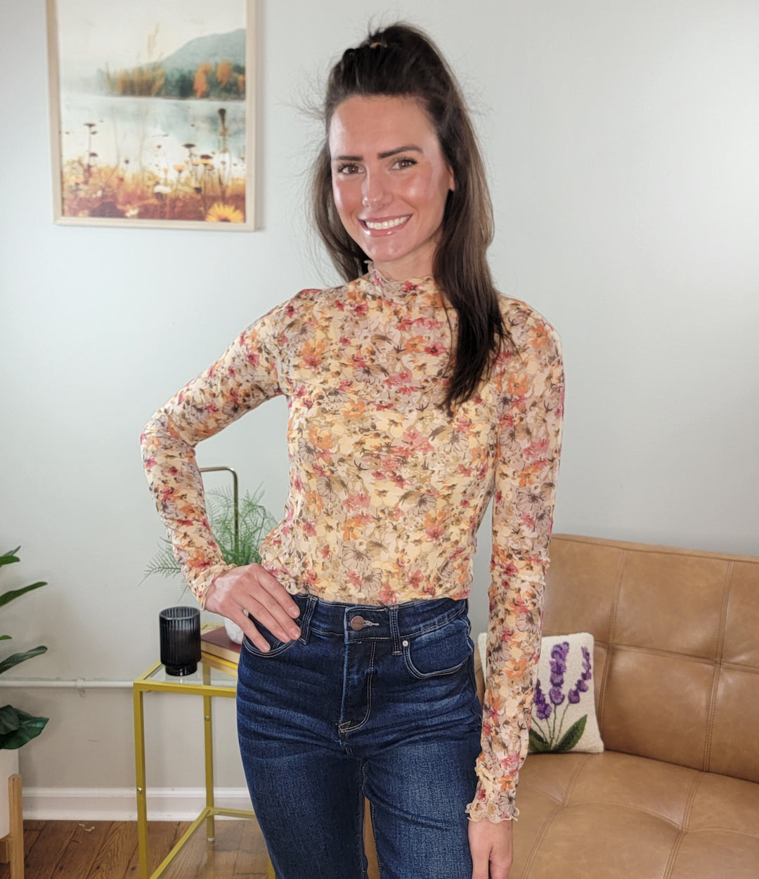 A woman with long dark hair smiles in a cozy living room, wearing the Hem & Thread Tatum Floral Mesh Top layered over a mock neck with blue jeans, surrounded by a tan couch, plants, and wall art.