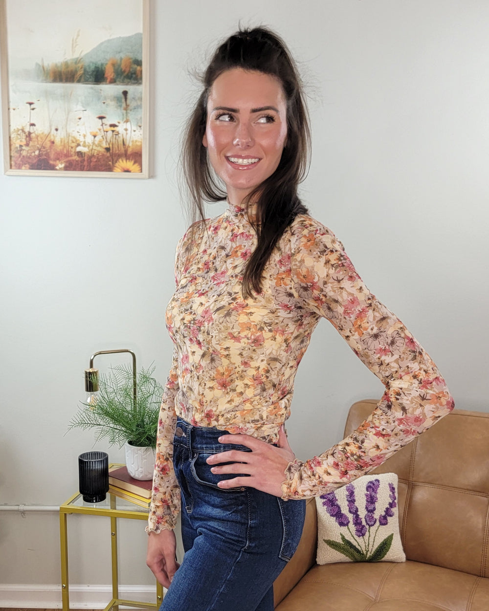 A woman with long brown hair wears the Hem & Thread Tatum Floral Mesh Top and blue jeans, smiling in a living room with a beige couch, lavender pillow, and wall art in the background.