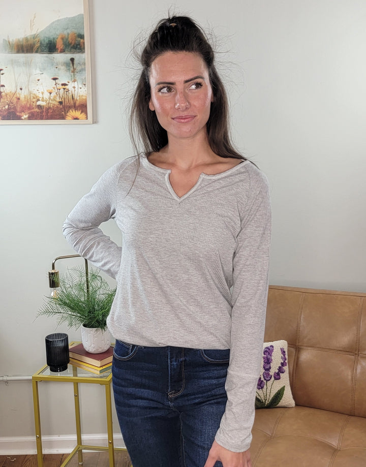 A woman with long dark hair wears the Lovely Melody Kyssa Long Sleeve Thermal V-Neck Top in heather grey and dark jeans, standing indoors by a tan sofa and a yellow side table with plants and books. A framed nature painting is displayed behind her.