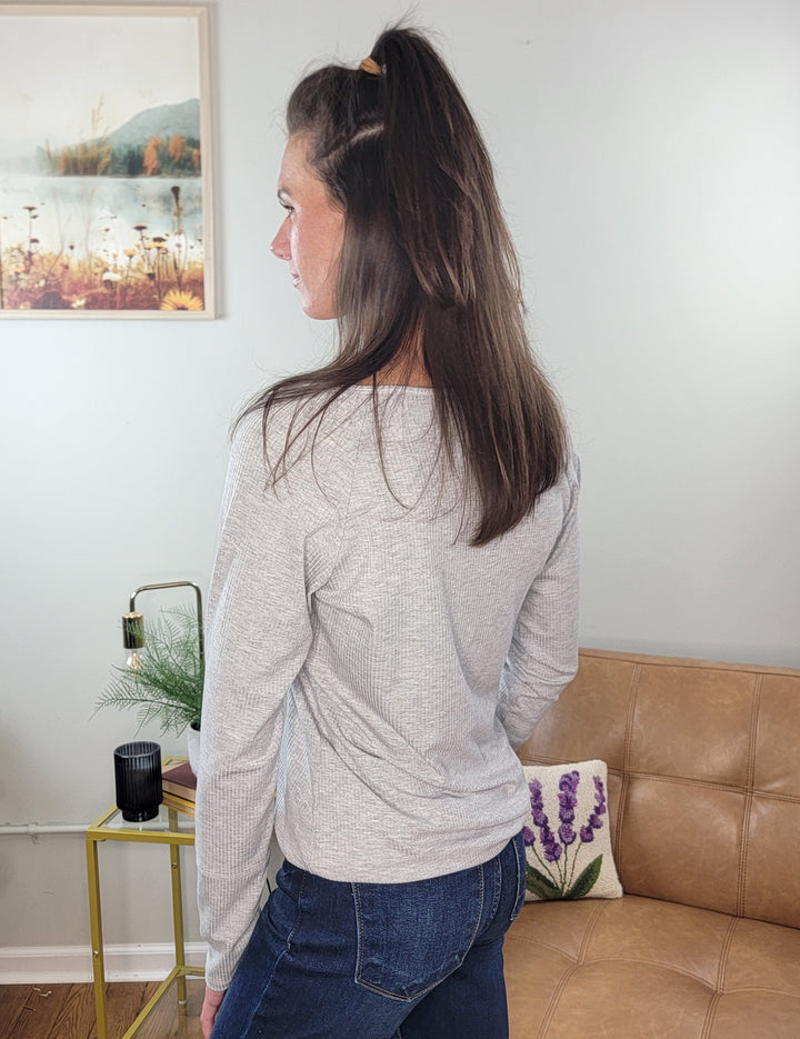 A woman with long brown hair stands indoors, facing away from the camera, wearing a Lovely Melody Kyssa Long Sleeve Thermal V-Neck Top in heather grey and blue jeans. Behind her are a tan couch, side table, black lamp, and framed landscape picture.