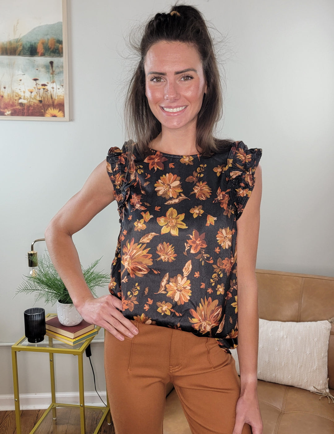 A woman with long brown hair smiles indoors, wearing the Doe & Rae Jenna Floral Ruffle Top paired with rust-colored pants. She stands by a tan couch and a small table—an ideal look to refresh your fall wardrobe.