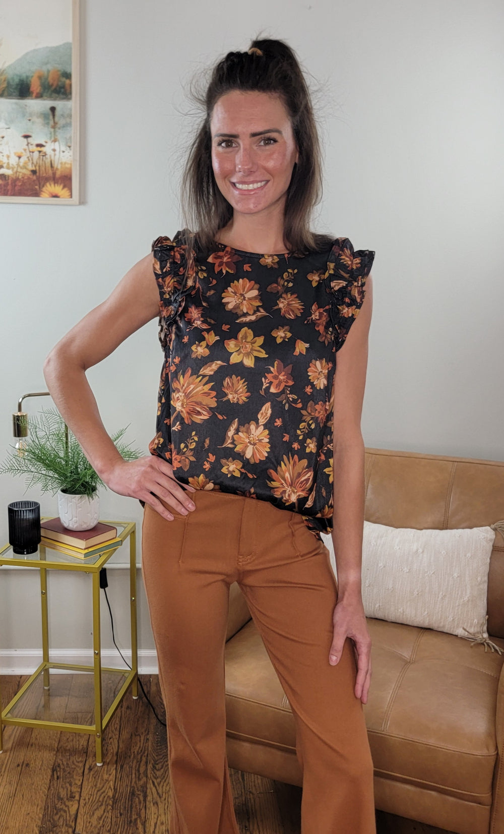 A woman with long brown hair stands indoors, smiling and wearing the Doe & Rae Jenna Floral Ruffle Top—an inspiring choice for your fall wardrobe. Behind her are a tan couch, a yellow table with a plant, and wall art.
