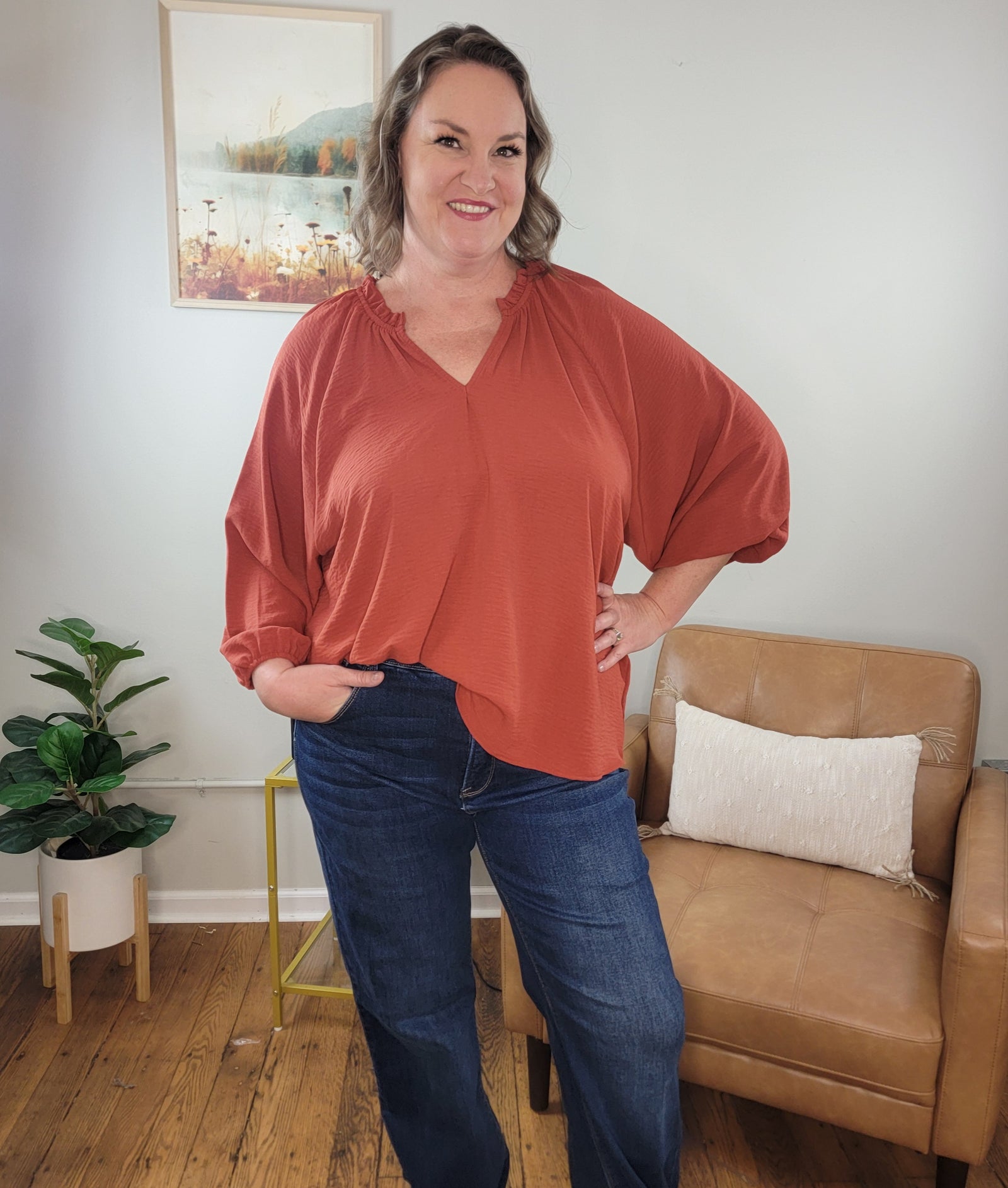 A woman with shoulder-length brown hair smiles indoors, wearing the ee:some Macie Long Sleeve, Light Weight Dressy Top with V-Neck in rust and blue jeans. One hand is in her pocket; behind her are a tan armchair, potted plant, and framed landscape.