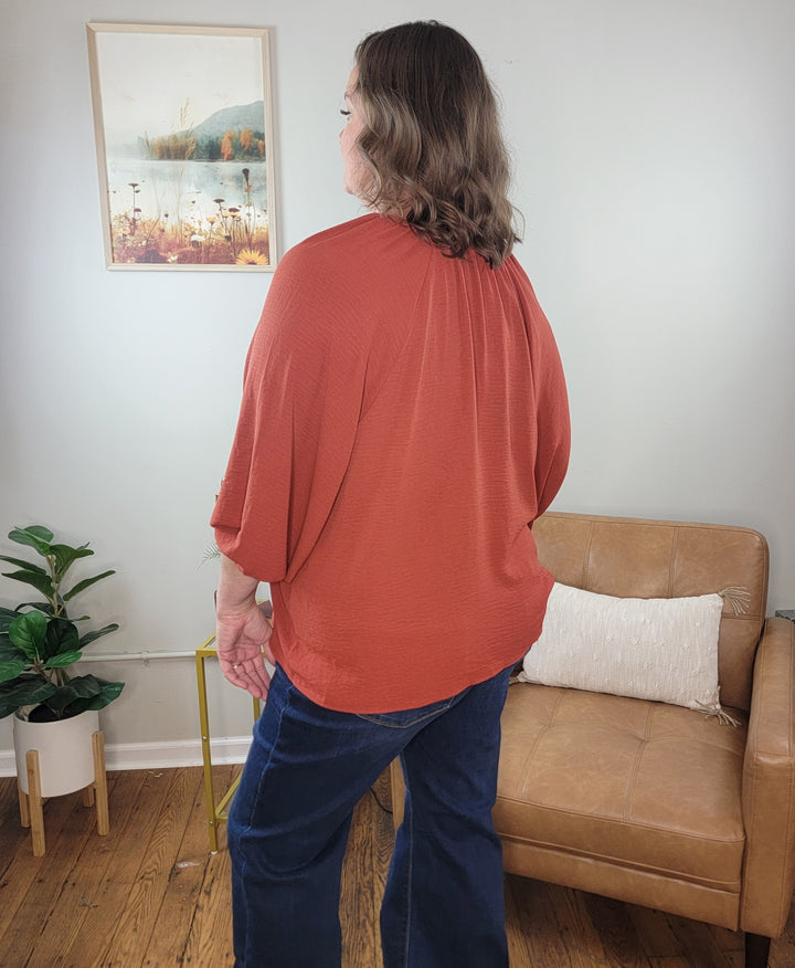 A person with wavy brown hair wears the ee:some Macie Top—a loose-fitting rust-colored v-neck—paired with blue jeans. They stand in a living room featuring a tan chair, wooden floor, plant, and framed nature photo.