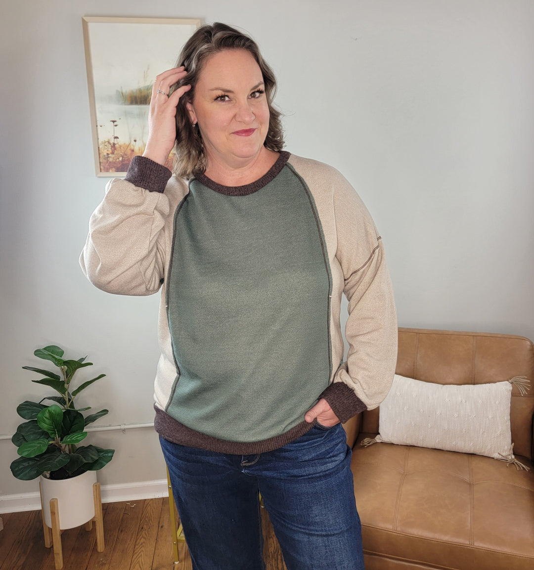 A woman wearing a Lovely Melody Michelle Color Block Knit Sweatshirt featuring a sage green front panel, beige sleeves, and dark brown ribbed trim at the collar, cuffs, and hem.