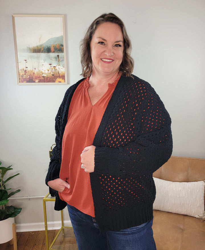 A smiling woman with wavy brown hair wears the Margot Open Front, Open Knit Cardigan by ee:some over a burnt orange blouse. She stands in a cozy living room with a tan sofa, plant, and framed nature print behind her.